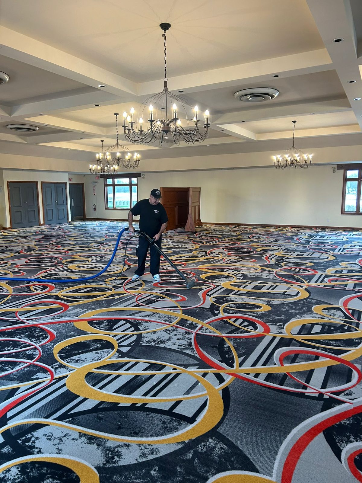 Technician steam cleaning the patterned carpet of a large banquet hall under chandeliers, hose running back toward the door